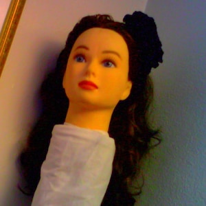 May include: A mannequin with long black hair and a black flower headband. The mannequin is wearing a white shirt.