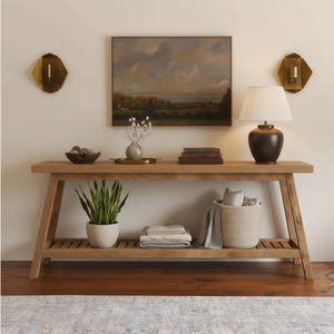 May include: A light wood console table with a landscape painting above it. A lamp with a white shade sits on the right. A snake plant, books, and decorative items are on the table. A woven basket and folded blankets are on the lower shelf.