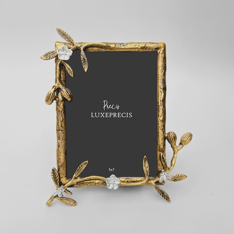 Peut inclure: Un cadre photo rectangulaire dor&eacute; avec une finition textur&eacute;e, orn&eacute; d'&eacute;l&eacute;ments floraux et de feuilles. Le cadre porte le texte "Precis LUXEPRECIS" et mesure 12,7 x 17,8 cm.