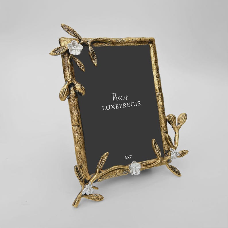 Peut inclure: Un cadre photo en m&eacute;tal dor&eacute; avec une ouverture rectangulaire. Le cadre est orn&eacute; de motifs floraux et de feuilles, ainsi que de petites fleurs blanches. Le cadre mesure 12,7 x 17,8 cm et porte le texte "LUXEPRECIS".