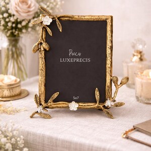 Peut inclure: Un cadre photo dor&eacute; avec un int&eacute;rieur noir, orn&eacute; d'&eacute;l&eacute;ments floraux et de feuilles. Le cadre rectangulaire porte le texte "Precis LUXEPRECIS" et "5x7". Le cadre est pos&eacute; sur une surface blanche.