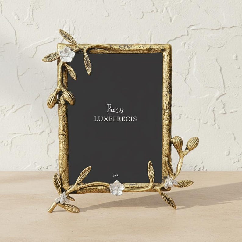 Peut inclure: Un cadre photo en m&eacute;tal dor&eacute; avec une finition textur&eacute;e, orn&eacute; de motifs floraux et de feuilles. Le cadre est rectangulaire et porte les inscriptions "Precis LUXEPRECIS" et "5x7".