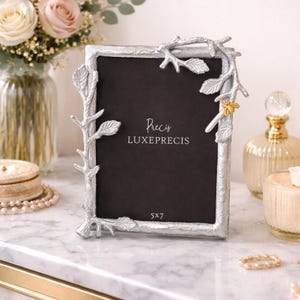 May include: Silver-coloured photo frame with a botanical design. The frame features branches and leaves, with a small gold-coloured bee accent. The frame is designed to hold a 12.7 x 17.8 cm photo. The text "Precy LUXEPRECIS" is printed on the black background.