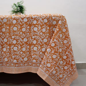 Op de afbeelding: Een rechthoekig tafelkleed met een mosterdgele achtergrond en een herhalende witte bloemen- en granaatappelprint. De stof heeft een decoratieve rand met een kleiner bloemmotief en een afwerking met witte en gele strepen.