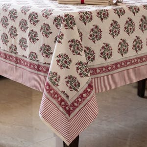 Può includere: Una tovaglia rettangolare con base color bianco sporco e un motivo ripetuto di disegni floreali rosso scuro e verdi. I bordi sono rifiniti con un'ampia fascia decorativa a motivi floreali e un motivo a sottili righe verticali.