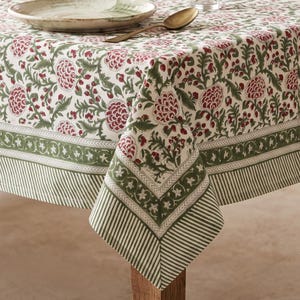 Può includere: Una tovaglia con sfondo color crema e un motivo floreale ripetuto in rosso scuro e verde oliva. Il tessuto presenta un bordo ampio con una finitura a righe verdi e bianche e una fascia decorativa con piccoli motivi floreali bianchi su verde.