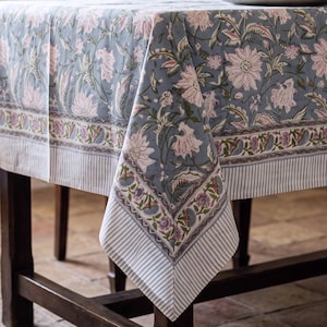 May include: A rectangular tablecloth featuring a dusty blue background with a repeating floral pattern in soft pink and green. The fabric has a wide decorative border with smaller floral motifs and a thin white and grey striped edge finish.