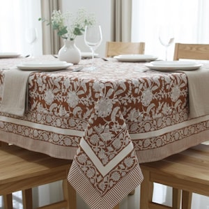 May include: A rectangular tablecloth featuring a rust-colored background with a white floral and botanical print. The fabric has a wide border with a striped pattern along the edges. The tablecloth is draped over a table set with white plates.