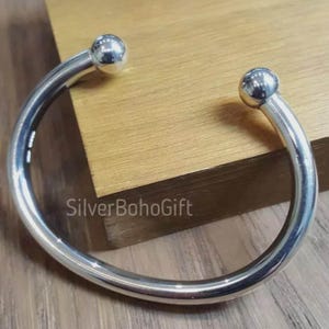 May include: A silver cuff bracelet with a rounded design. The bracelet has a smooth, polished finish and features a small, spherical accent at each end. The text "SilverBohoGift" is visible on the surface. The bracelet is displayed on a wooden surface.