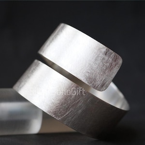Op de afbeelding: Een zilveren ring met een geborstelde metalen textuur. De ring heeft een uniek, open ontwerp met een opening in het midden. De donkere achtergrond benadrukt de glans van het metaal. De tekst "SohoGift" is zichtbaar.