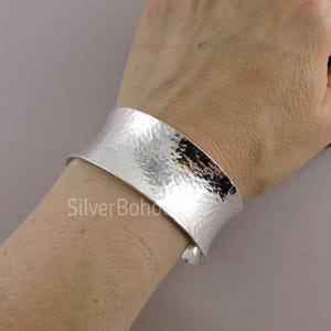 May include: A wide, hammered silver cuff bracelet. The bracelet has a textured surface and a curved design. The text "SilverBoho" is visible on the image. This piece of jewellery is a stylish accessory.
