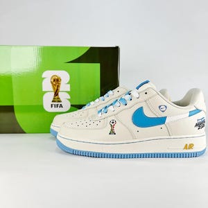 Puede incluir: Zapatillas de color crema con detalles en azul claro, incluyendo el logo de Nike, los cordones y la suela. Las zapatillas presentan detalles de la FIFA y Argentina 2026, con un pequeño emblema de trofeo. Una caja verde con la marca FIFA 26 está en el fondo.