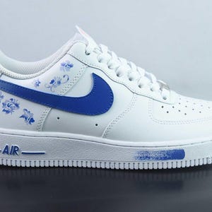 May include: White leather trainer with blue floral patterns and a blue swoosh. The sole is white with blue detailing and the word "AIR" in blue. A stylish athletic shoe.