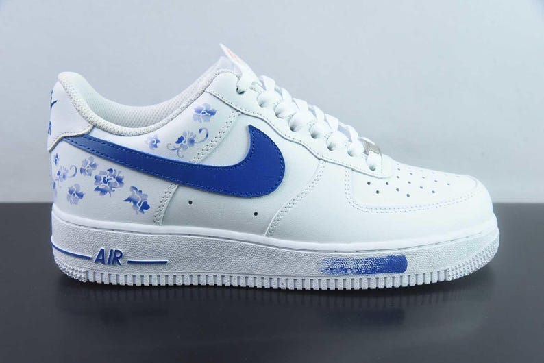 May include: White leather trainer with blue floral patterns and a blue swoosh. The sole is white with blue detailing and the word "AIR" in blue. A stylish athletic shoe.