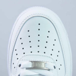 May include: Close-up of a white trainer's toe box. The shoe has a perforated design and a small metal tag with "AF1". The trainer is made of smooth, white leather. The design is classic.