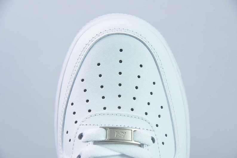 May include: Close-up of a white trainer's toe box. The shoe has a perforated design and a small metal tag with "AF1". The trainer is made of smooth, white leather. The design is classic.