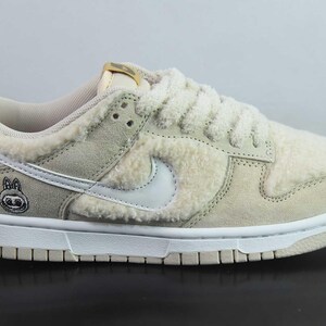 Peut inclure: Baskets beiges et blanches avec une tige textur&eacute;e et duveteuse. La chaussure pr&eacute;sente un logo Swoosh blanc, des lacets blancs et une semelle blanche. Un petit personnage de dessin anim&eacute; est sur le c&ocirc;t&eacute; de la chaussure.