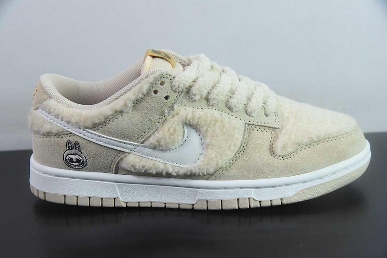 Peut inclure: Baskets beiges et blanches avec une tige textur&eacute;e et duveteuse. La chaussure pr&eacute;sente un logo Swoosh blanc, des lacets blancs et une semelle blanche. Un petit personnage de dessin anim&eacute; est sur le c&ocirc;t&eacute; de la chaussure.