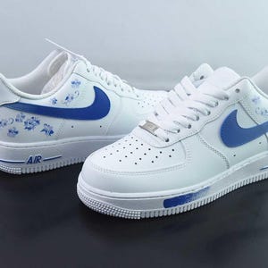 May include: White leather trainers with blue detailing, featuring the Nike swoosh and floral designs. The trainers have white laces, a white sole, and the word "AIR" in blue on the side.