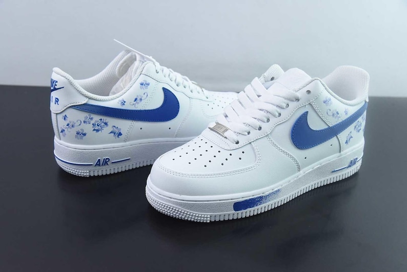 May include: White leather trainers with blue detailing, featuring the Nike swoosh and floral designs. The trainers have white laces, a white sole, and the word "AIR" in blue on the side.