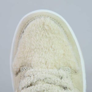 Peut inclure: Gros plan d'une basket beige avec une tige textur&eacute;e et duveteuse. La chaussure est dot&eacute;e d'une semelle et de lacets blancs. Le design est simple, mettant l'accent sur la mati&egrave;re douce et moelleuse.