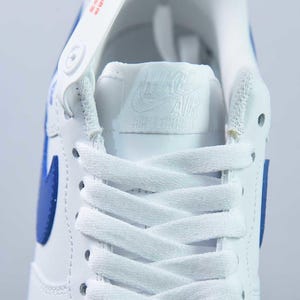 May include: Close-up of a white and blue athletic shoe. The shoe features white laces, a blue swoosh, and a white tongue with the brand logo. The shoe is made of leather and has a classic design.