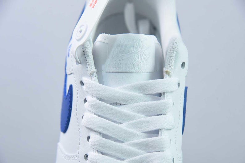 May include: Close-up of a white and blue athletic shoe. The shoe features white laces, a blue swoosh, and a white tongue with the brand logo. The shoe is made of leather and has a classic design.