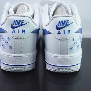 May include: Pair of white Nike Air Force 1 trainers with blue detailing. The heel displays the Nike logo and "AIR" in blue, with a floral pattern. The soles have a blue stripe.