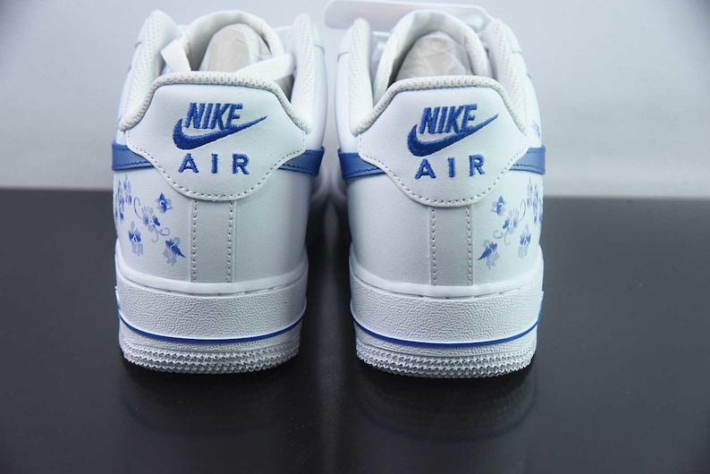 May include: Pair of white Nike Air Force 1 trainers with blue detailing. The heel displays the Nike logo and "AIR" in blue, with a floral pattern. The soles have a blue stripe.