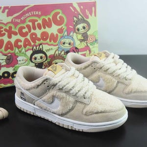 Peut inclure: Des baskets Nike Dunk Low beiges et blanches avec une texture duveteuse, des semelles blanches et un logo Nike blanc. Les baskets sont &agrave; c&ocirc;t&eacute; d'une bo&icirc;te avec le texte "Exciting Macaron" et des personnages de dessins anim&eacute;s. Une paire de lacets beiges est &eacute;galement pr&eacute;sente.