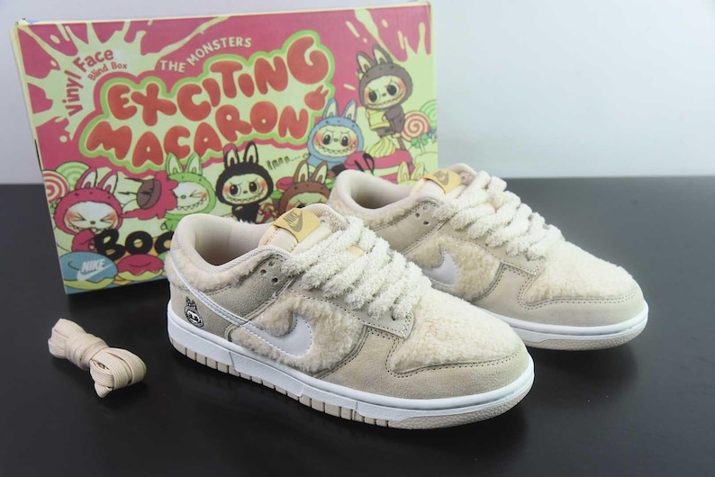 Peut inclure: Des baskets Nike Dunk Low beiges et blanches avec une texture duveteuse, des semelles blanches et un logo Nike blanc. Les baskets sont &agrave; c&ocirc;t&eacute; d'une bo&icirc;te avec le texte "Exciting Macaron" et des personnages de dessins anim&eacute;s. Une paire de lacets beiges est &eacute;galement pr&eacute;sente.