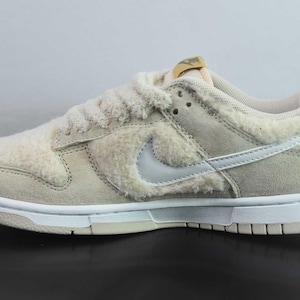Peut inclure: Une basket Nike Dunk Low beige et blanche. La chaussure pr&eacute;sente un mat&eacute;riau duveteux et textur&eacute; sur le dessus, avec un logo Swoosh blanc et une semelle blanche. La languette a une &eacute;tiquette dor&eacute;e. La basket est con&ccedil;ue pour un usage d&eacute;contract&eacute;.