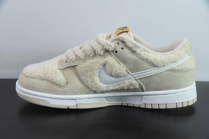Peut inclure: Une basket Nike Dunk Low beige et blanche. La chaussure pr&eacute;sente un mat&eacute;riau duveteux et textur&eacute; sur le dessus, avec un logo Swoosh blanc et une semelle blanche. La languette a une &eacute;tiquette dor&eacute;e. La basket est con&ccedil;ue pour un usage d&eacute;contract&eacute;.