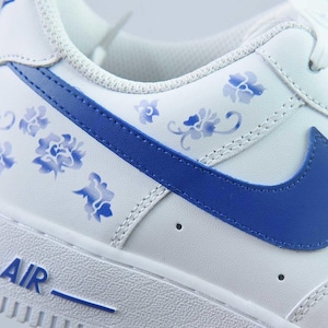 May include: White leather trainer with blue floral detailing and a blue Nike swoosh. The word "AIR" is printed in blue on the white sole. The shoe features a classic design with a modern floral twist.