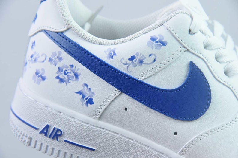 May include: White leather trainer with blue floral detailing and a blue Nike swoosh. The word "AIR" is printed in blue on the white sole. The shoe features a classic design with a modern floral twist.