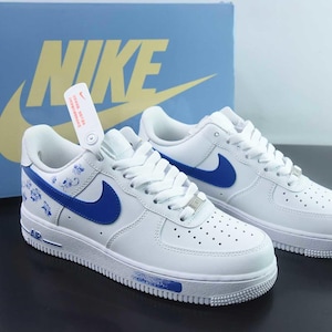 May include: White leather trainers with blue detailing, including the Nike swoosh and floral patterns. The shoes are shown with a blue box featuring the Nike logo. A tag hangs from one shoe.