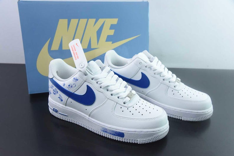 May include: White leather trainers with blue detailing, including the Nike swoosh and floral patterns. The shoes are shown with a blue box featuring the Nike logo. A tag hangs from one shoe.