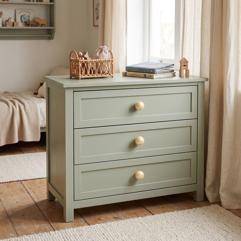 May include: A sage green wooden dresser featuring three horizontal drawers, each equipped with a single round, light-colored wooden knob. The piece has a clean, rectangular frame and sits on four short, sturdy legs, providing a stable storage solution.