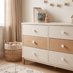 May include: A six-drawer dresser featuring a two-tone design with cream-colored and light wood-grain drawer fronts, accented by round white knobs. The dresser stands on short wooden legs and is positioned next to a woven wicker basket.