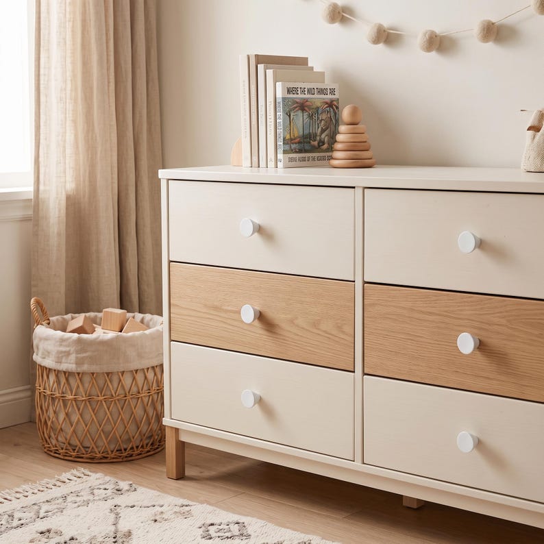 May include: A six-drawer dresser featuring a two-tone design with cream-colored and light wood-grain drawer fronts, accented by round white knobs. The dresser stands on short wooden legs and is positioned next to a woven wicker basket.