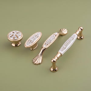May include: A collection of four gold-toned cabinet hardware pieces, including one round knob, one oval knob, and two handles. Each piece features a white inlay decorated with small, clear, sparkling crystals set into the metallic surface.