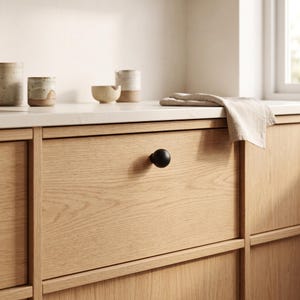 Op de afbeelding: Een matzwarte, bolvormige kastknop gecentreerd op een lichte eikenhouten ladefront. De lade heeft een strak, vlak ontwerp met zichtbare houtnerfpatronen, aangevuld met een neutraal gekleurde linnen doek die er vlakbij ligt.