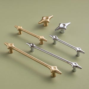 May include: A collection of cabinet hardware including two long matte gold drawer pulls, one long polished silver drawer pull, one small matte gold knob, and one small polished silver knob, all featuring a distinct four-pointed star shape.