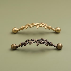 May include: Two metal cabinet pulls featuring a vine and leaf design. One pull has a brushed gold finish, while the other has a dark bronze finish. Both pulls have a curved center section with detailed leaves and a spherical knob at each end.