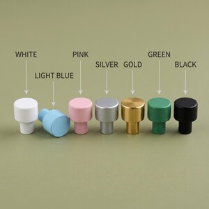 Op de afbeelding: Een set van zeven cilindrische kastknoppen in verschillende kleuren, waaronder wit, lichtblauw, roze, zilver, goud, groen en zwart. Elke knop is voorzien van de bijbehorende kleurnaam erboven, wat de beschikbare kleuropties aangeeft.