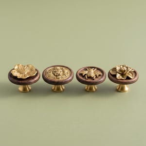 May include: Four circular cabinet knobs featuring dark brown wooden bases and gold-toned metal tops. The designs include a flower, a lion head, a bee, and a multi-petaled blossom, each mounted on a small gold-toned metal pedestal base.