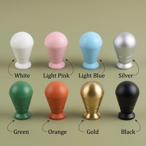 Op de afbeelding: Acht kastknoppen gerangschikt in twee rijen, elk met een afgeronde bovenkant en een taps toelopende basis met twee horizontale groeven. De kleuren zijn wit, lichtroze, lichtblauw, zilver, groen, oranje, goud en zwart, met labels eronder.