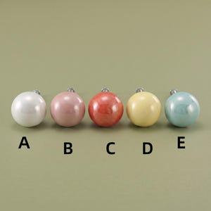 May include: Five round cabinet knobs arranged in a horizontal row, labeled A through E. The knobs are white, dusty pink, coral, pale yellow, and light blue, each featuring a smooth, glossy finish and a small metal mounting screw at the back.