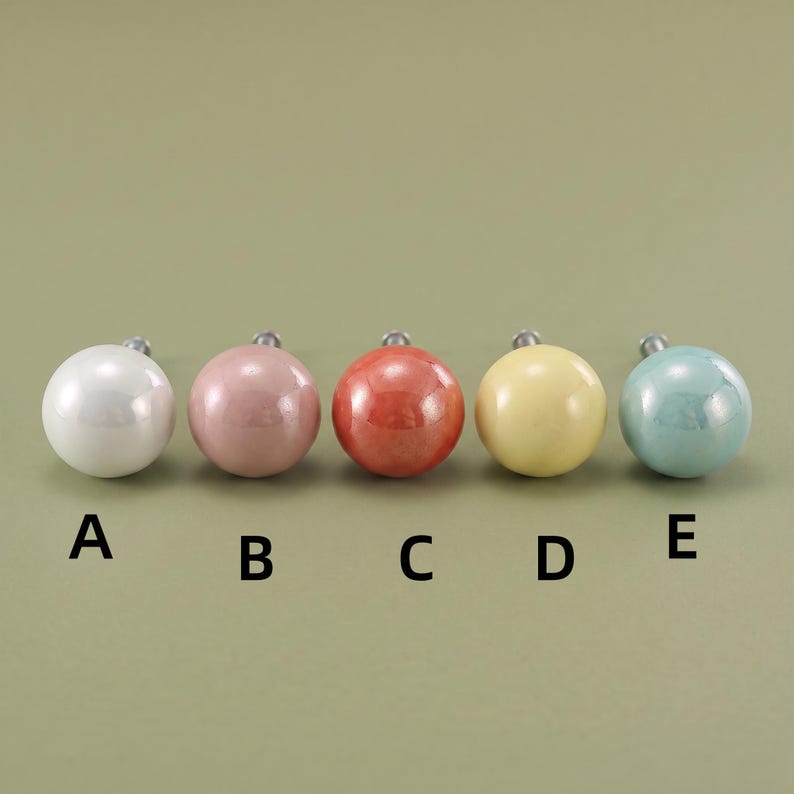 May include: Five round cabinet knobs arranged in a horizontal row, labeled A through E. The knobs are white, dusty pink, coral, pale yellow, and light blue, each featuring a smooth, glossy finish and a small metal mounting screw at the back.