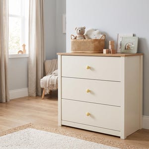 May include: A three-drawer dresser with an off-white frame and a light wood-toned top surface. Each drawer features a single round, light-colored wooden knob. The dresser is positioned against a wall, with a woven basket and books on top.